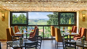 Un restaurante con vistas a ondulantes colinas y techo de madera. Las mesas están dispuestas con sillas y centros de mesa.