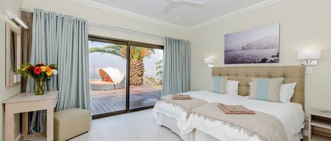 Habitación de hotel con cama, escritorio, silla, ventana con cortinas, ventilador de techo y un cuadro en la pared.