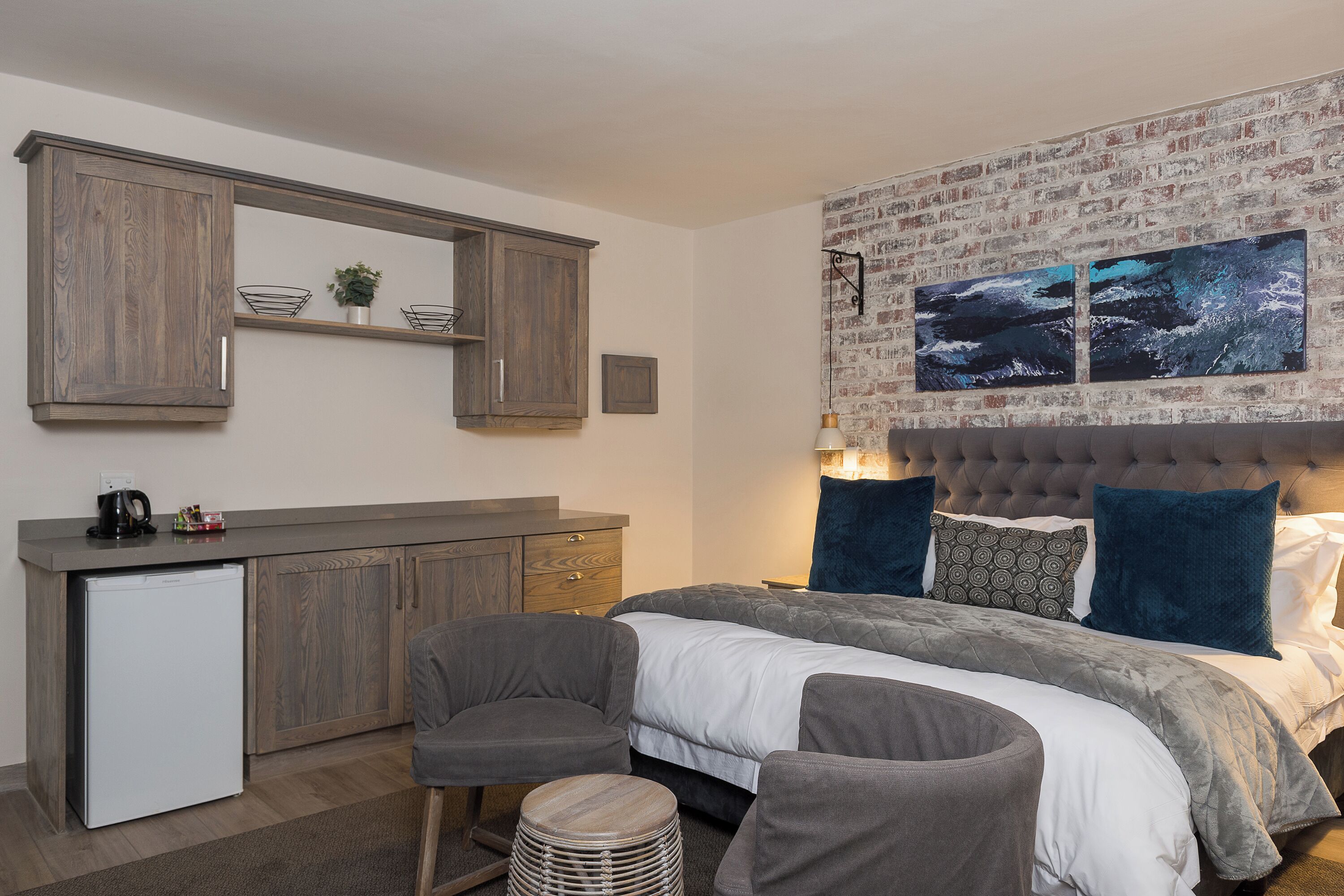 A compact bedroom with a bed, bedside tables, a small kitchenette, and a brick-patterned headboard.