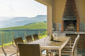 Un espacio de comedor al aire libre con vistas a colinas verdes, un horno de ladrillo y una barandilla de vidrio.