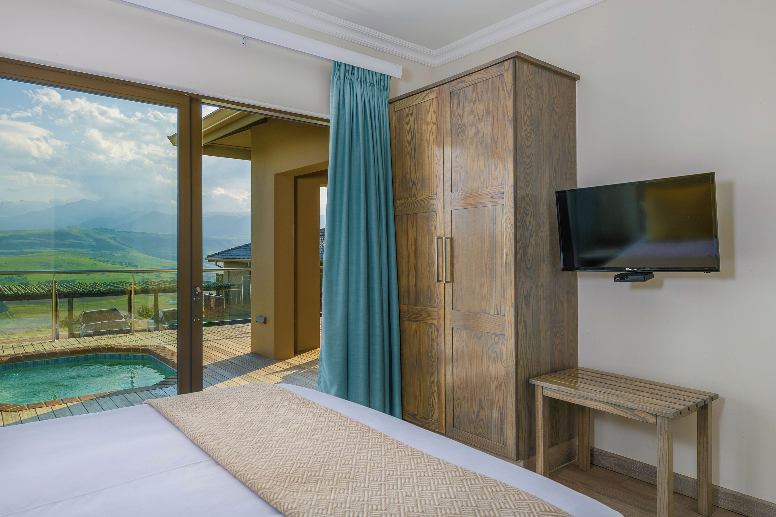 A bedroom with a large bed, a wooden wardrobe, a television, and a view of a swimming pool and landscape.