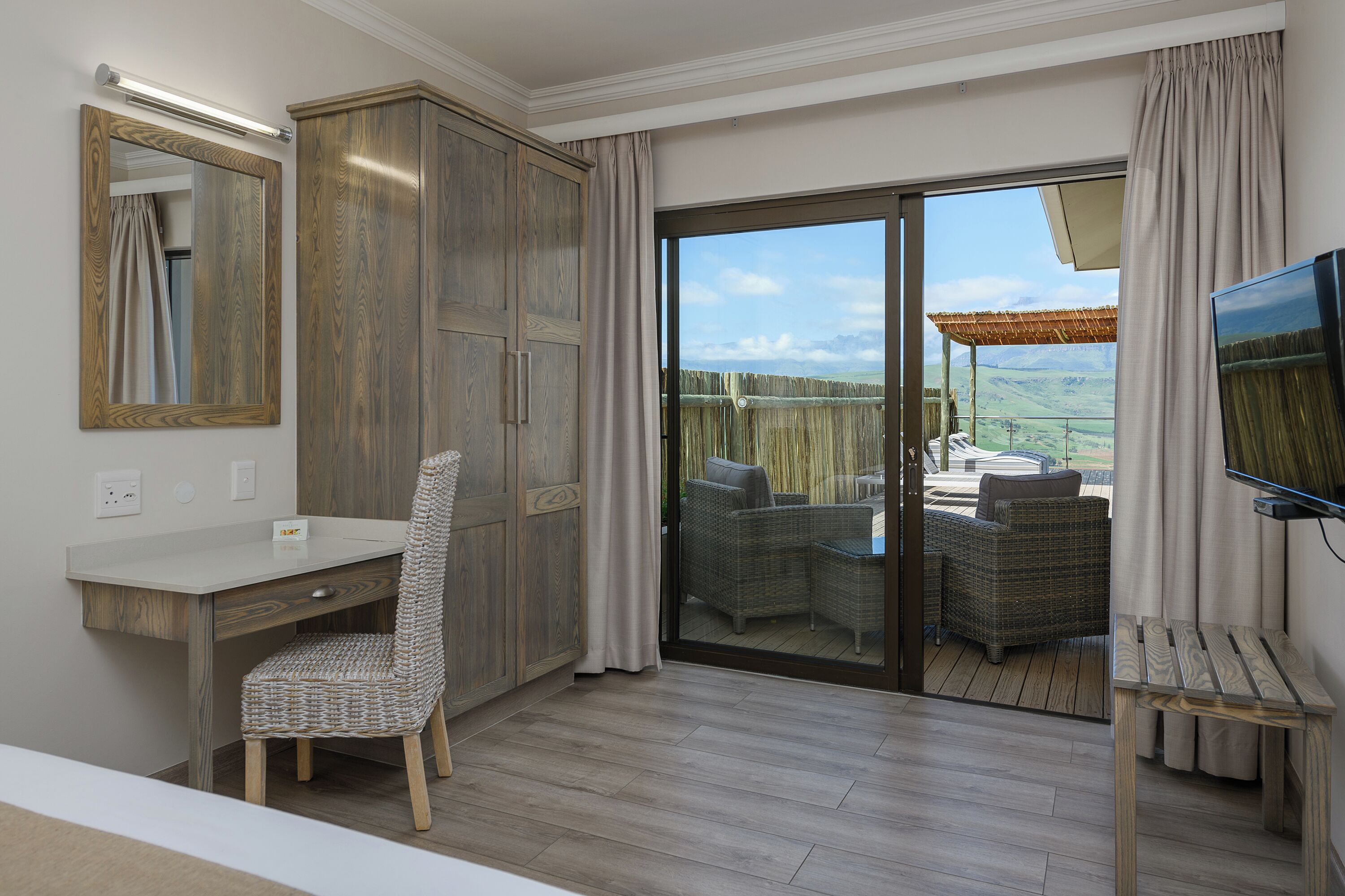 A room with a desk, chair, wardrobe, and a TV. It has a sliding glass door leading to an outdoor seating area with a view of the countryside.