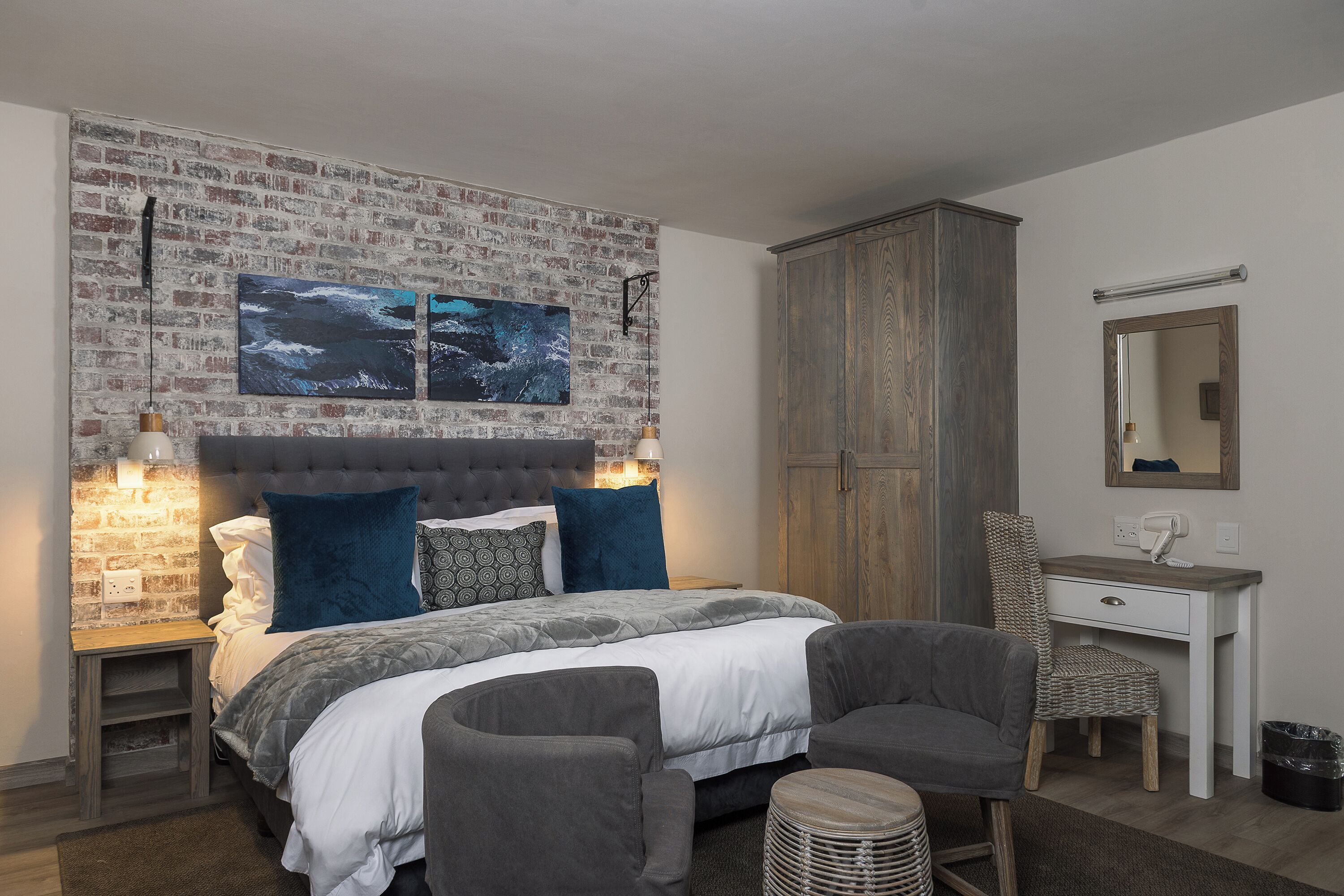 A bedroom with a brick-patterned headboard, a bed with grey and white bedding, two bedside lamps, a wooden wardrobe, a desk with a chair, and a mirror.