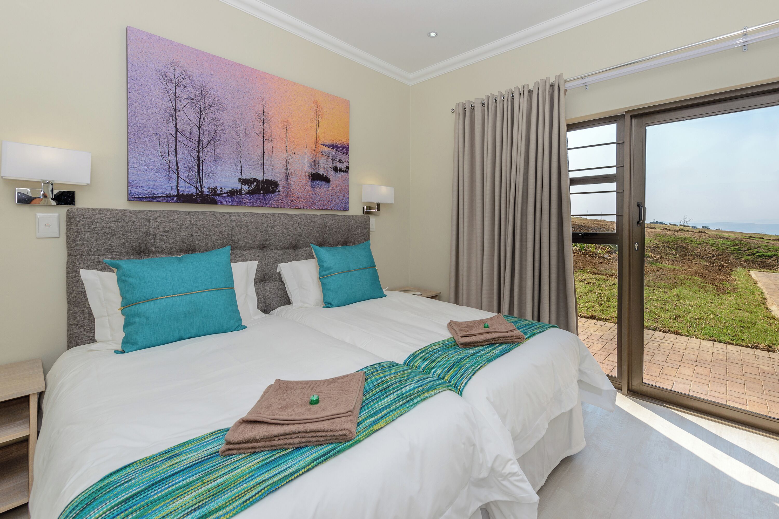 Un lit double avec une literie blanche et turquoise, un oreiller turquoise et une serviette marron. Un tableau représentant un coucher de soleil est accroché au mur, il y a une fenêtre avec des rideaux et une vue sur un espace verdoyant à l’extérieur.