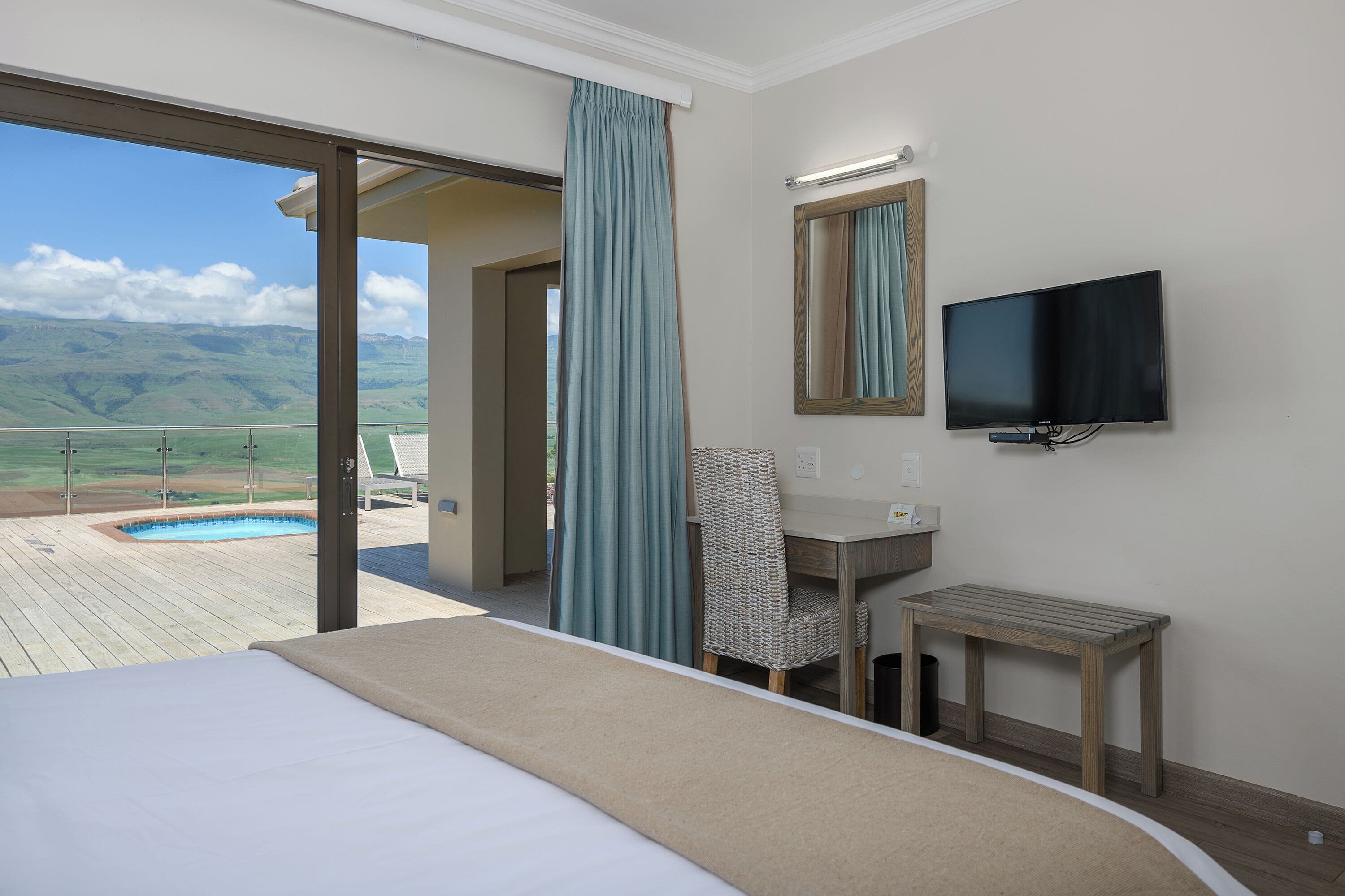 A hotel room with a bed, a desk, a chair, a TV, and a view of a pool and mountains.
