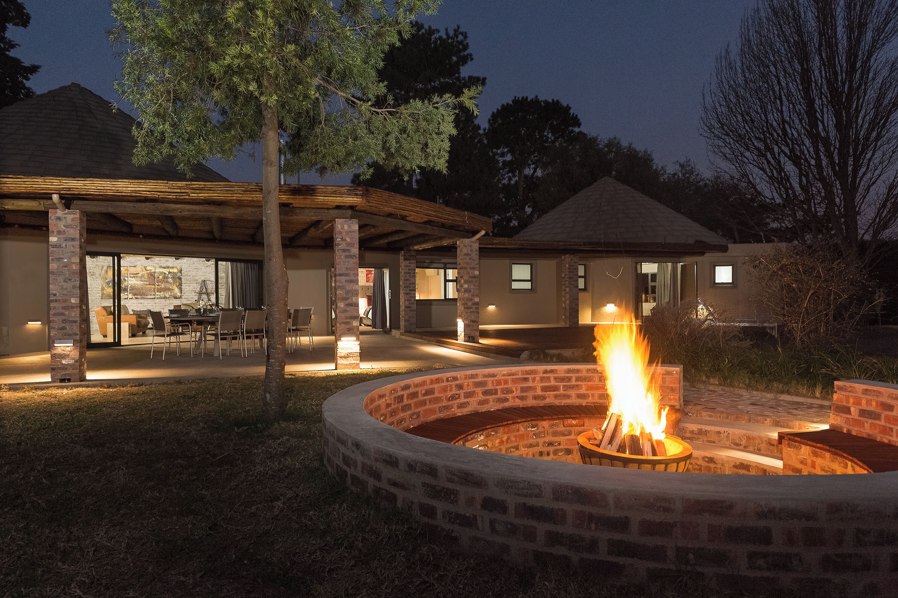 A circular brick fire pit with a fire, surrounded by a covered patio area with chairs and tables.