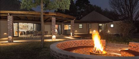 A circular brick fire pit with a fire, surrounded by a covered patio area with chairs and tables.
