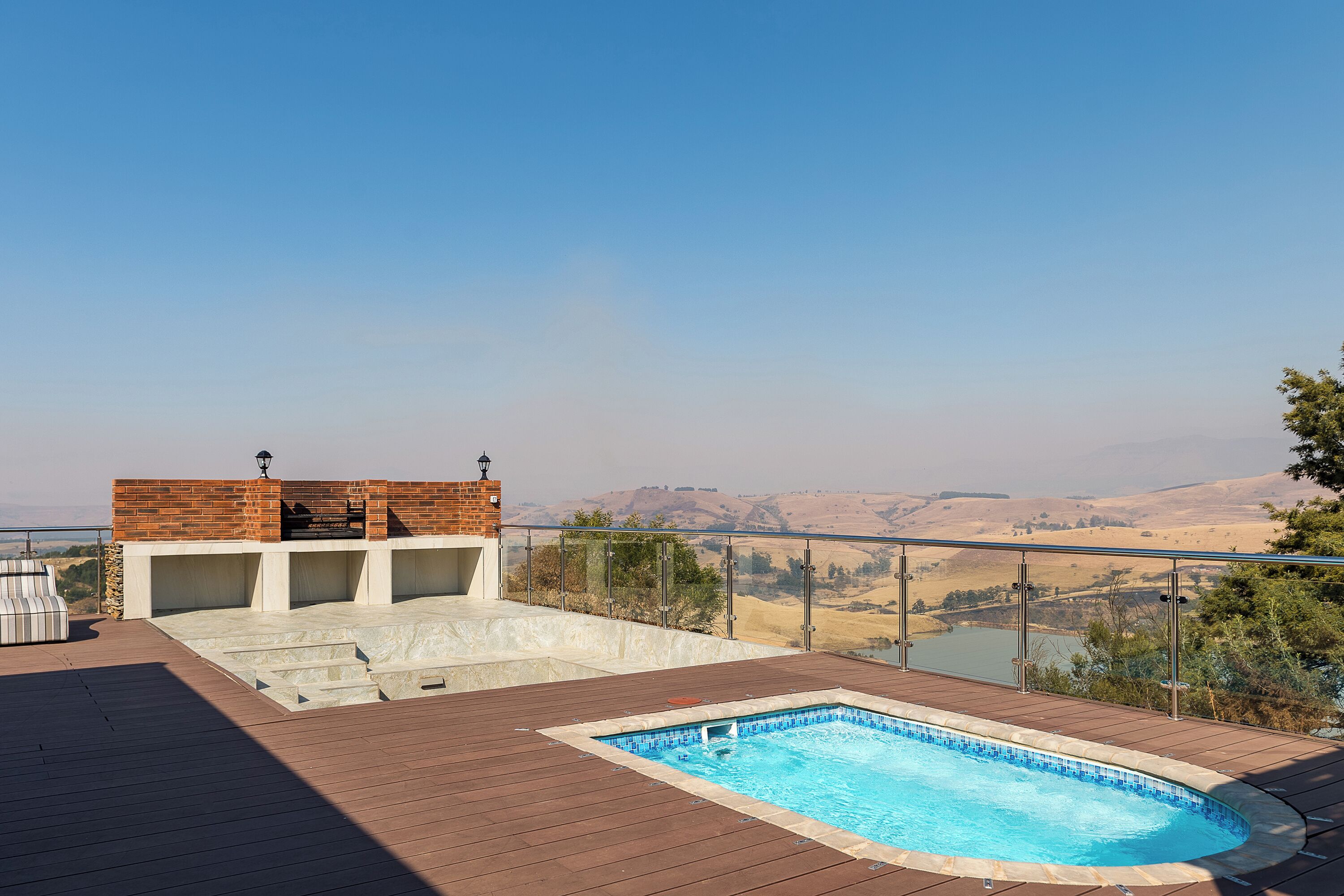 A swimming pool with a wooden deck, a barbecue area, and a view of hills.