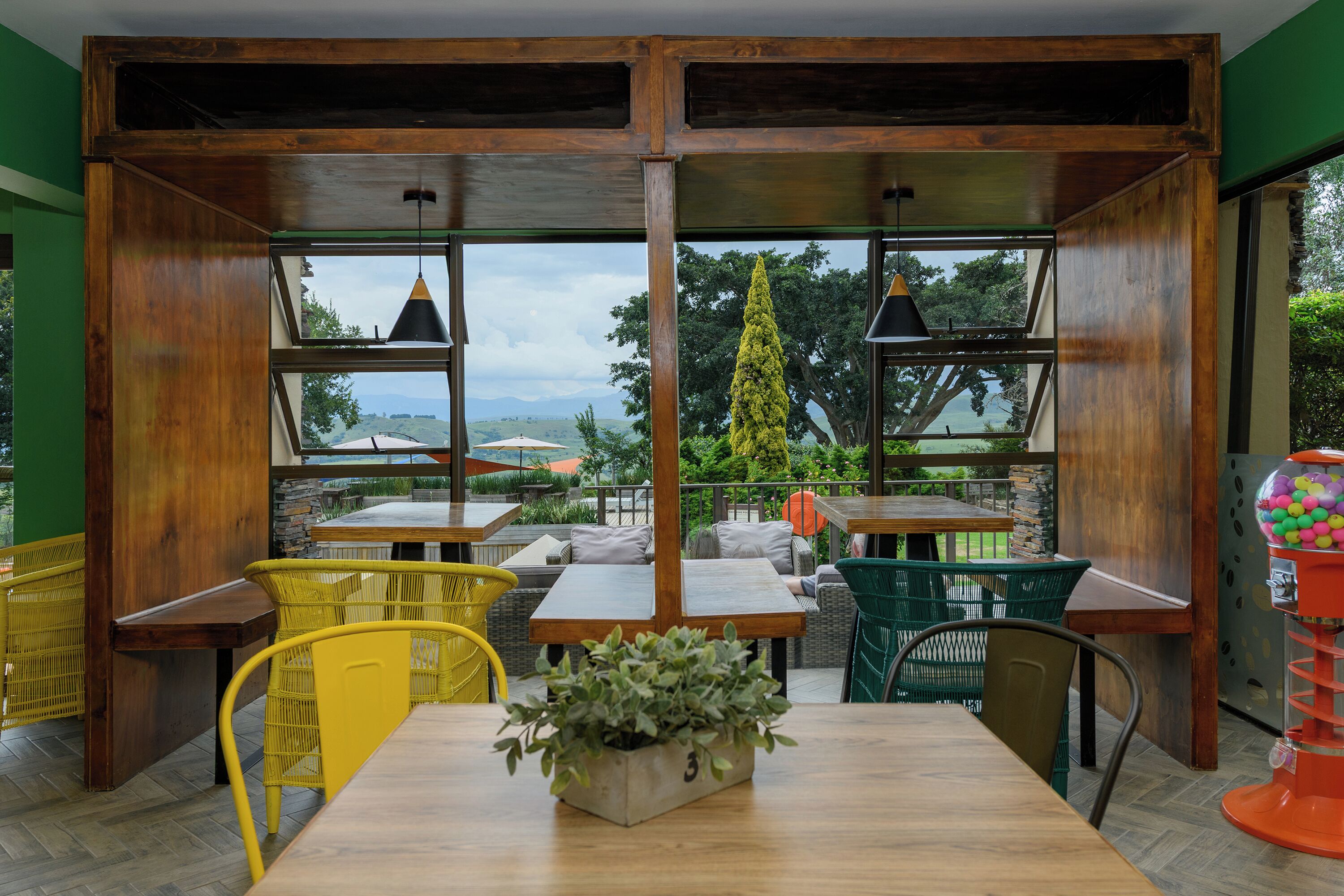 An outdoor seating area with wooden tables and chairs, a potted plant, and a view of greenery and hills.