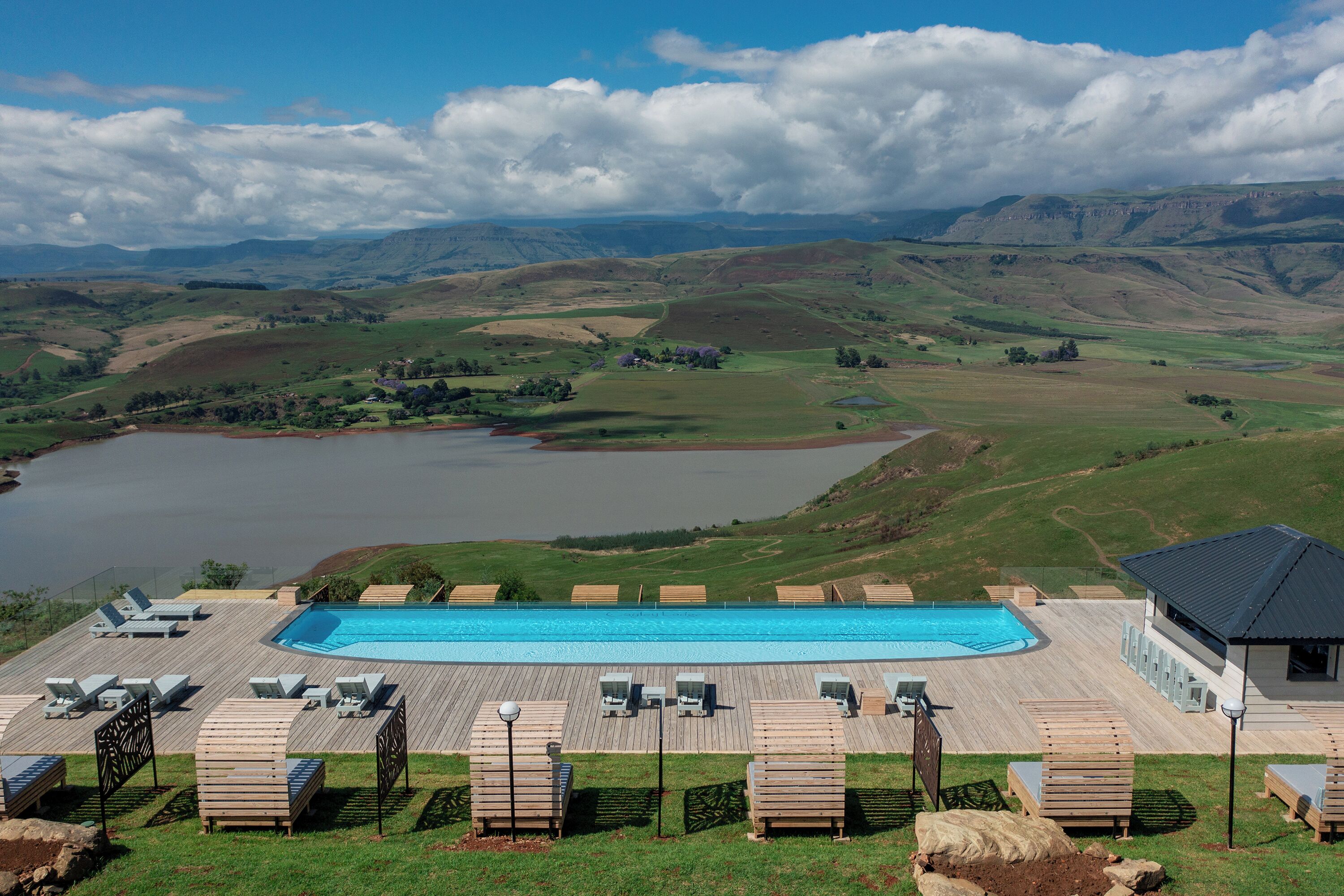 A pool area with lounge chairs, a wooden deck, and a view of hills and a lake.