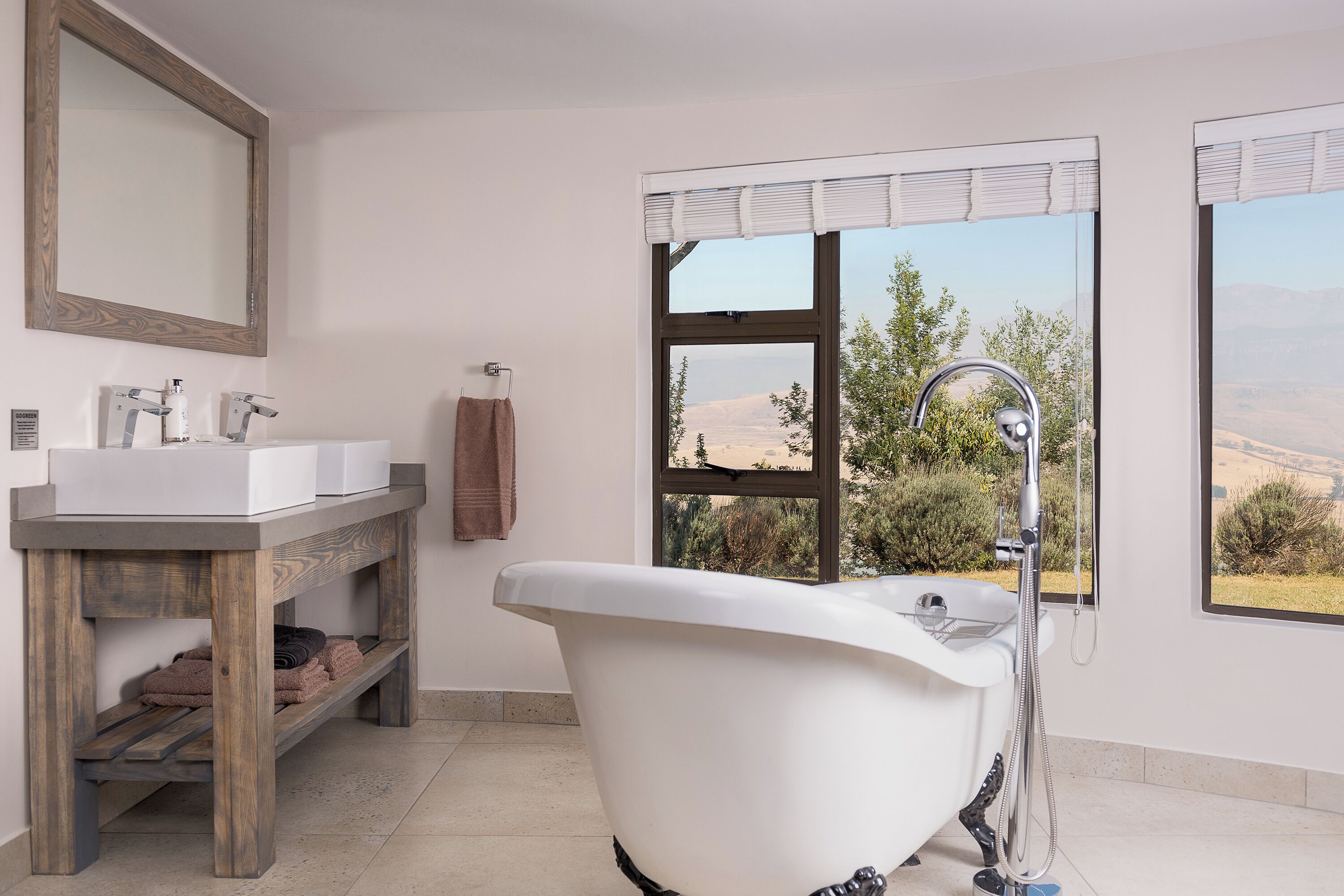 A bathroom with a bathtub, a wooden vanity, a large mirror, and a window with a view of the landscape.