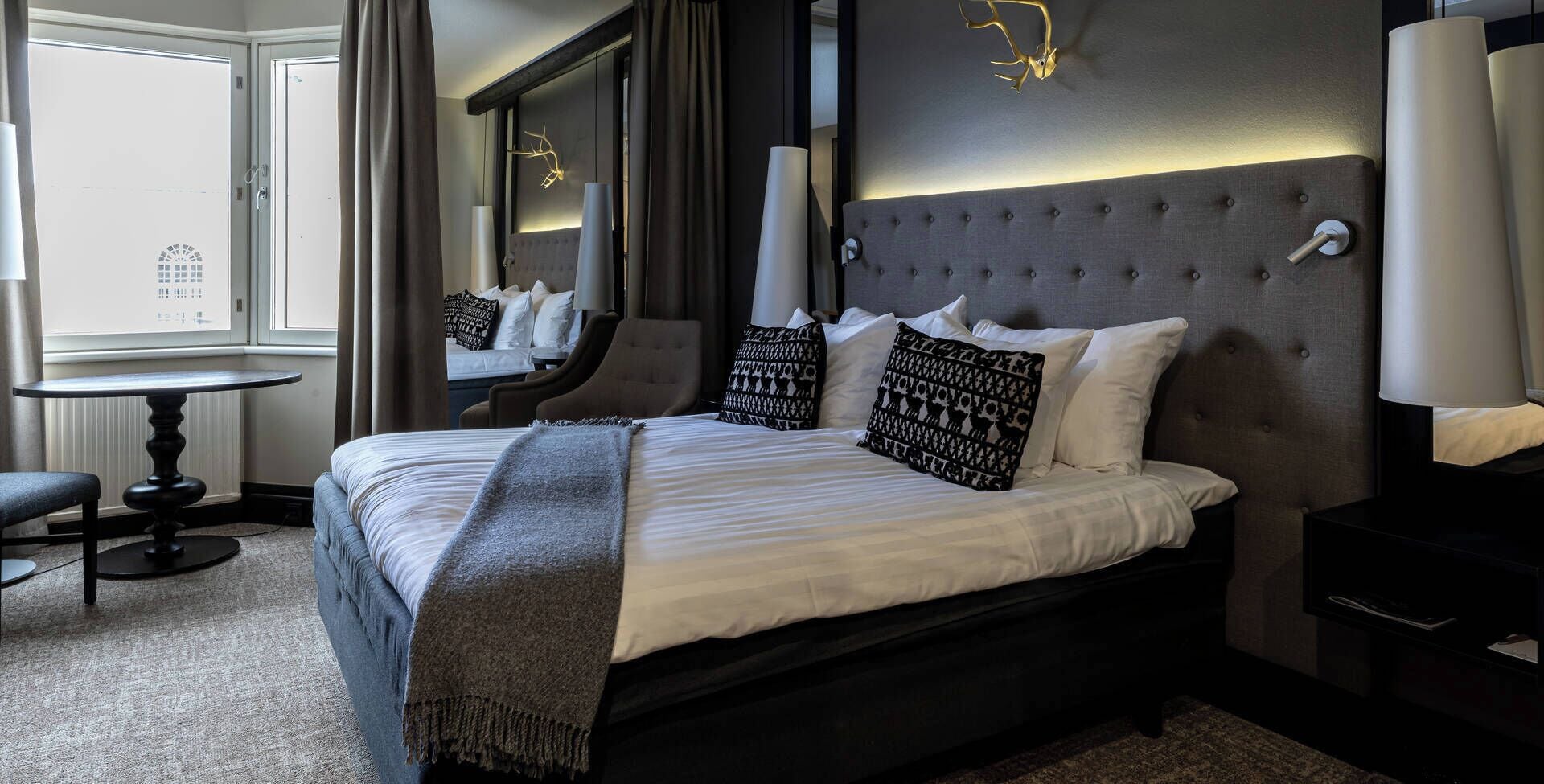 A hotel room with a large bed, a round table, a chair, a mirror, and a window with curtains.