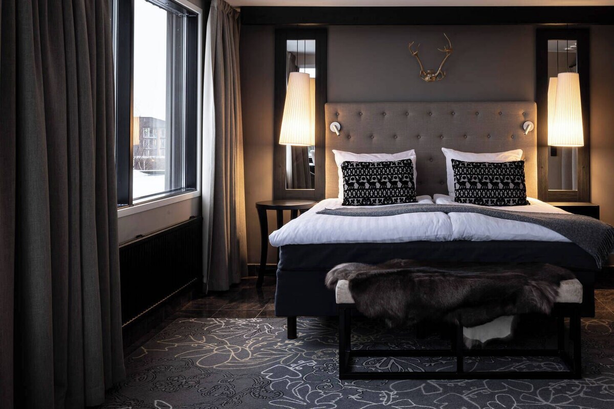 Une chambre d’hôtel avec un grand lit, deux lampes de chevet, une tête de cerf décorative et un banc recouvert de fourrure.