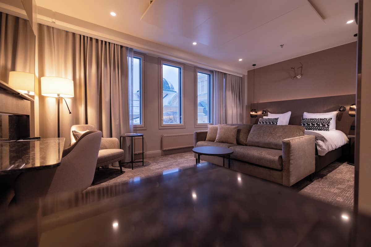 Une chambre d’hôtel moderne avec un canapé, une petite table ronde, un bureau avec une lampe et un lit agrémenté d’oreillers à motifs.