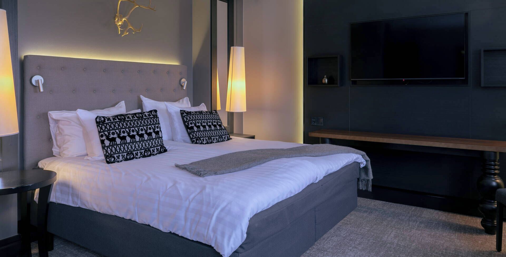Una cama bien tendida con ropa blanca y almohadas estampadas, una lámpara de noche, un televisor de pantalla plana montado en la pared y un banco de madera al lado de la cama.