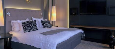 Una cama bien tendida con ropa blanca y almohadas estampadas, una lámpara de noche, un televisor de pantalla plana montado en la pared y un banco de madera al lado de la cama.