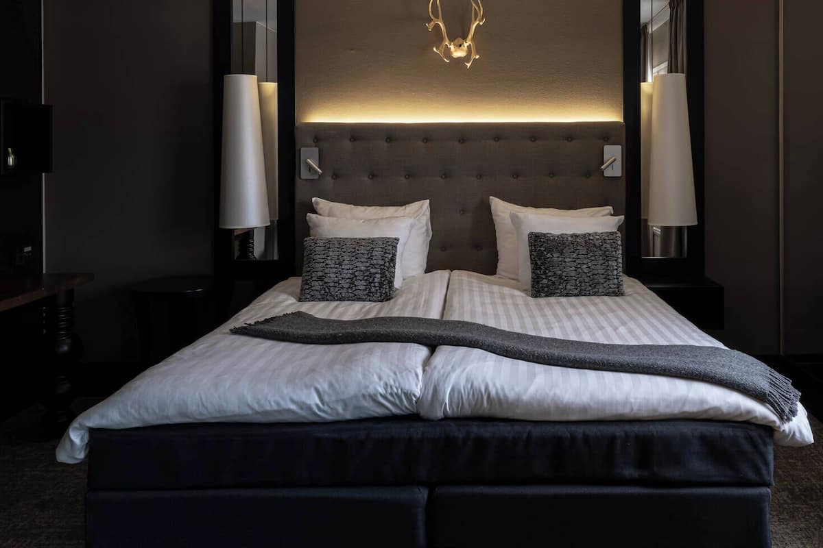 Un lit bien fait, avec une couverture grise, deux oreillers et une tête de lit agrémentée d’une lampe.