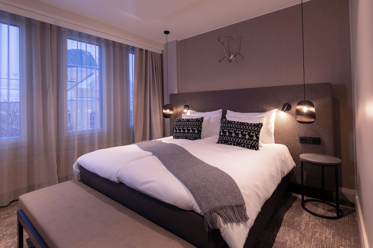 Une chambre d’hôtel moderne avec un grand lit, un banc au pied du lit, une petite table ronde et une vue sur un immeuble par la fenêtre.