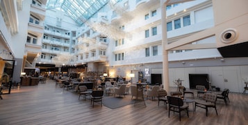 Um amplo hall de hotel com diversas áreas de estar, um bar e um teto alto com iluminação natural.