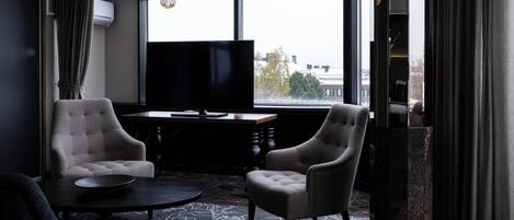 A room with two armchairs, a round table, a flat-screen TV, and a large window with a view of buildings and trees.