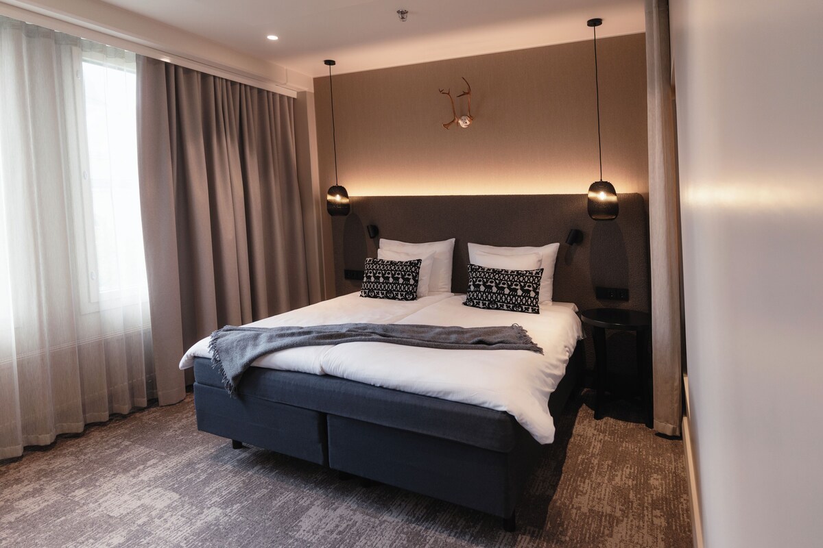 Une chambre d’hôtel moderne dotée d’un grand lit, d’une tête de lit avec éclairage intégré, d’une table de chevet et d’une grande fenêtre avec des rideaux.