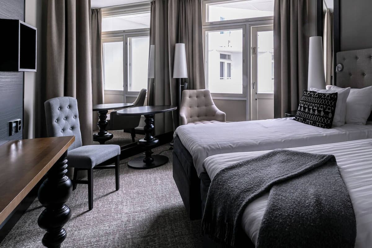 Une chambre d’hôtel avec deux lits, un bureau, une chaise et une vue sur les bâtiments, visible à travers de grandes fenêtres.