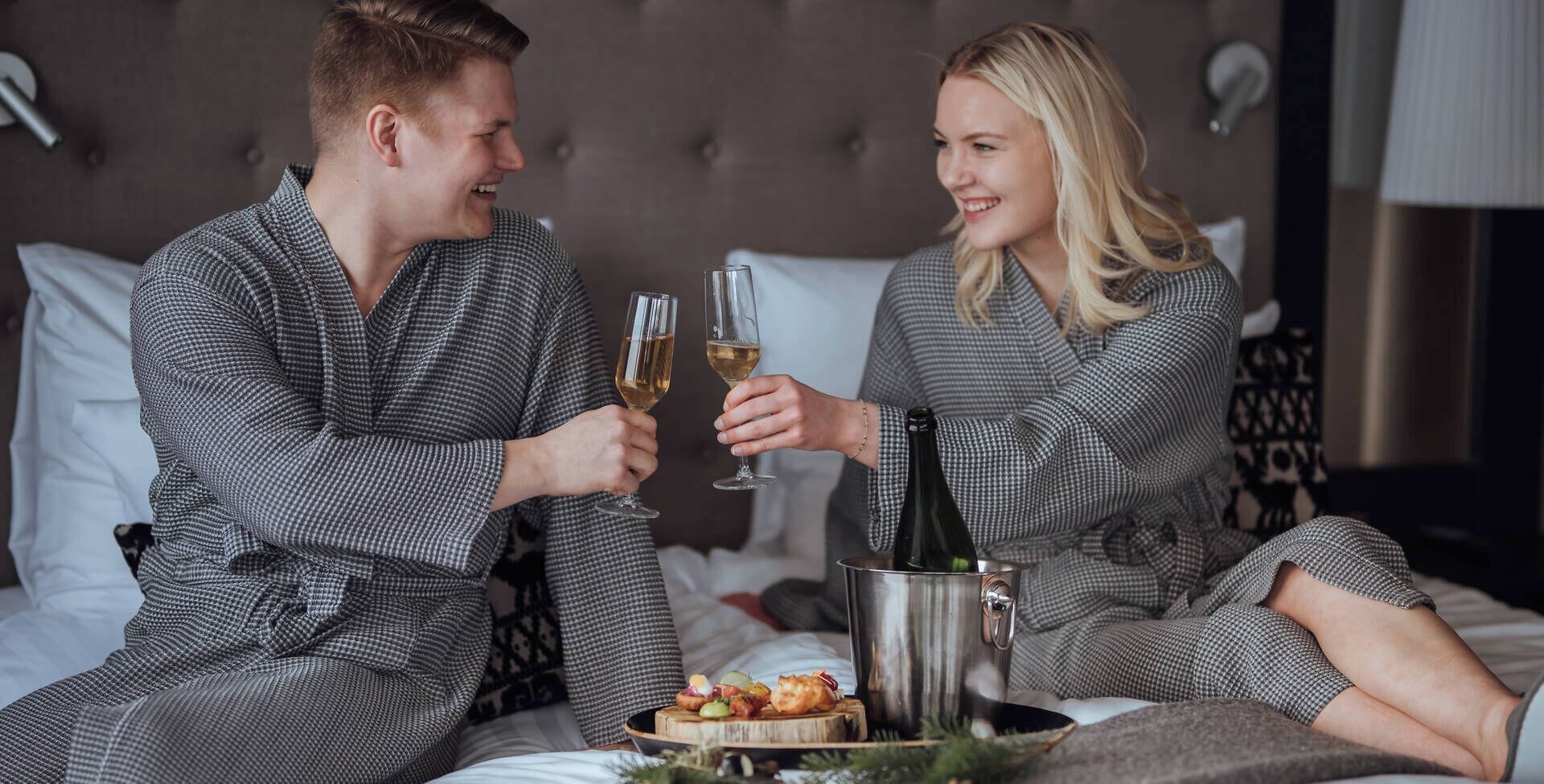 Una pareja con batas a juego sentada en una cama con una botella de champán y una bandeja de aperitivos.