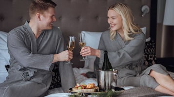 Una pareja con batas a juego sentada en una cama con una botella de champán y una bandeja de aperitivos.