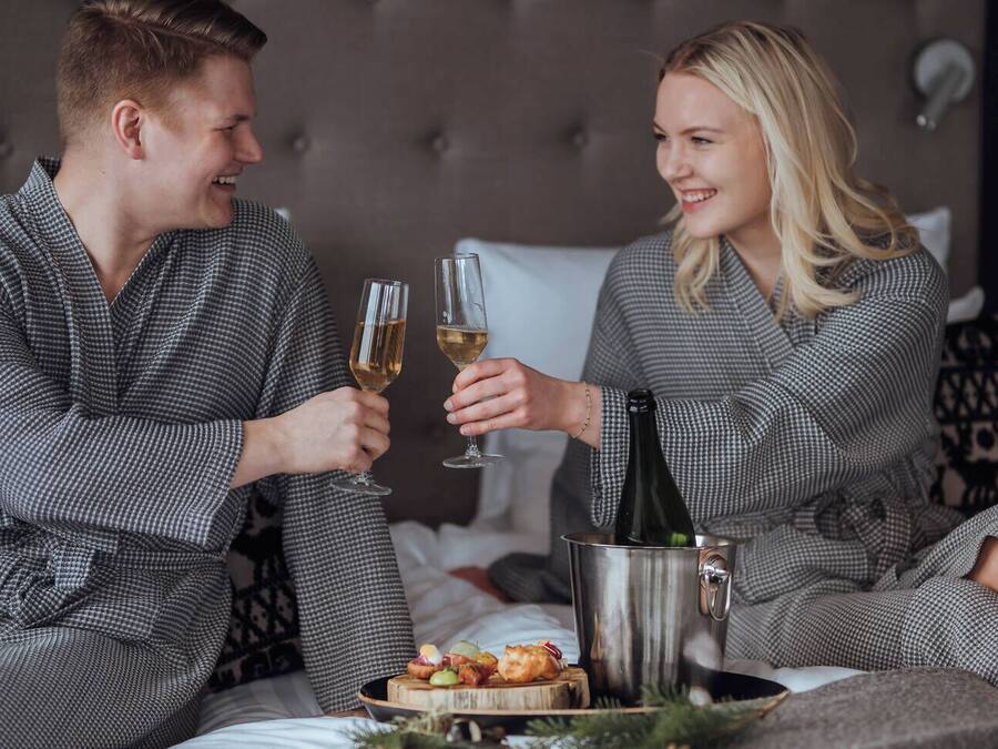 Una pareja con batas a juego sentada en una cama con una botella de champán y una bandeja de aperitivos.