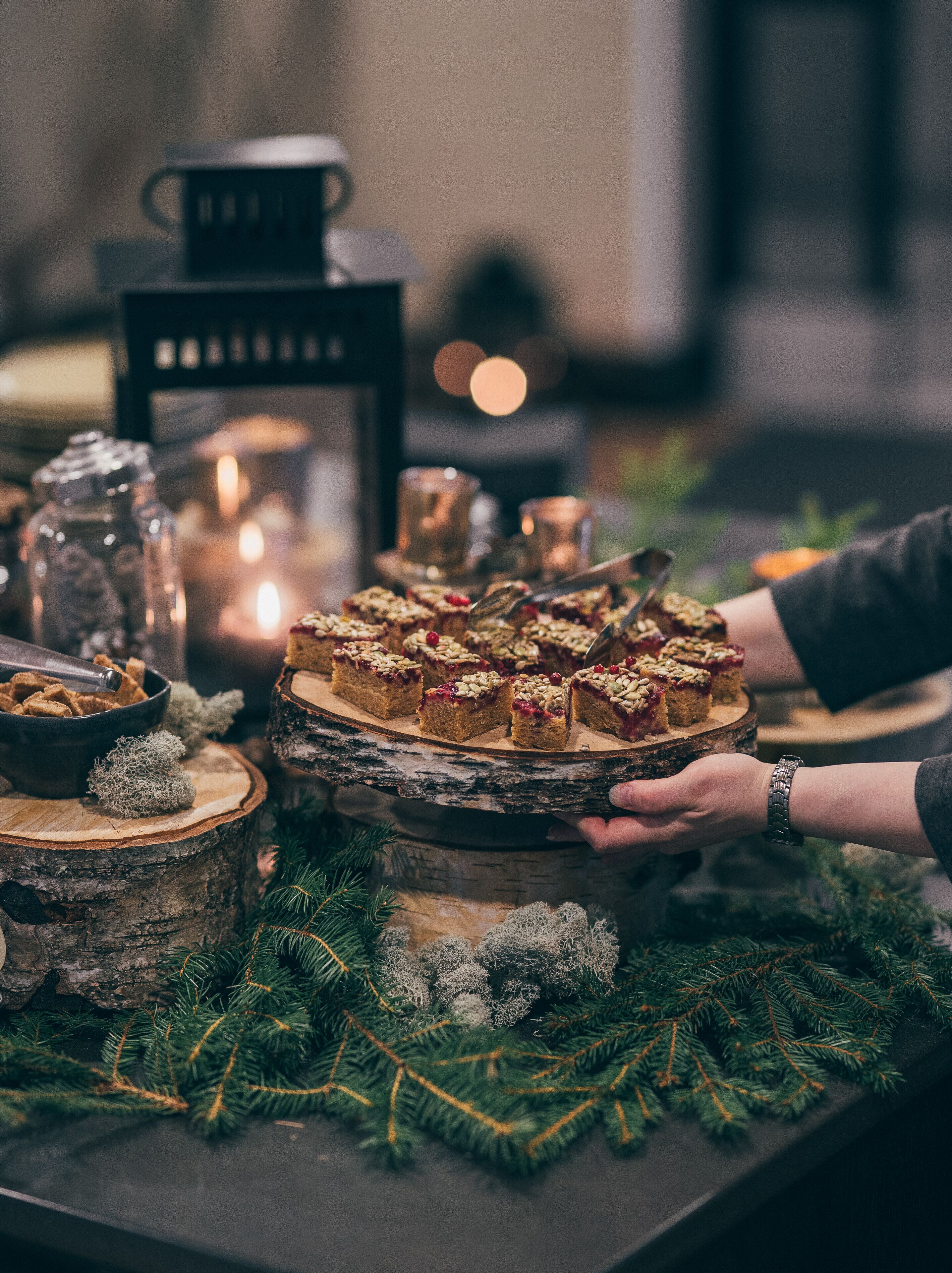 A table with a wooden platter holding dessert slices, surrounded by candles, a salt shaker, and Christmas decorations.