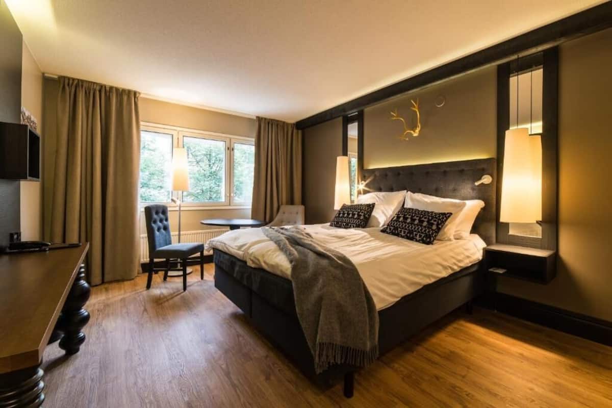 A modern hotel room with a large bed, a desk, and a chair. There is a window with curtains, a lamp, and a wall-mounted TV.