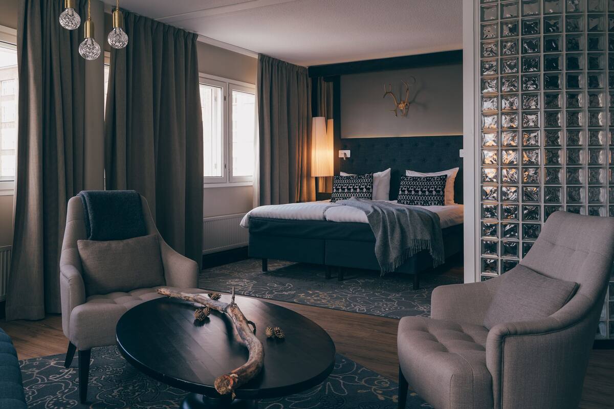 Une chambre d’hôtel comprenant un lit, deux fauteuils, une petite table basse ronde et une grande fenêtre avec des rideaux.