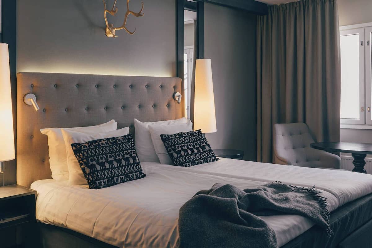 Une chambre d’hôtel avec un grand lit, deux lampes de chevet, une chaise, une petite table et une fenêtre avec des rideaux.