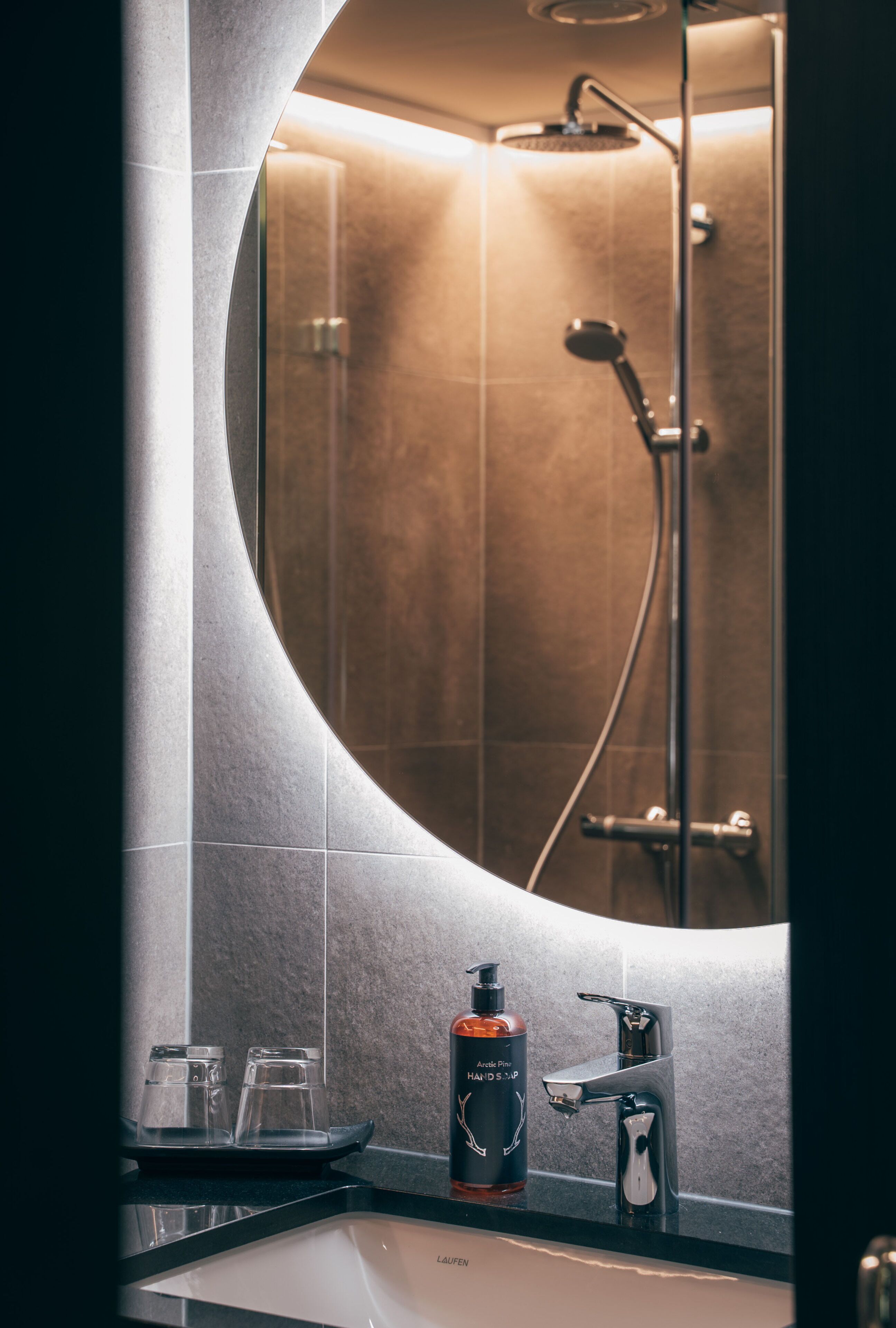 A bathroom with a round mirror, a sink, a shower, and a bottle of hand soap.