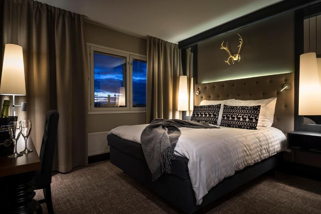 A modern hotel room with a large bed, a view of the cityscape through the window, and a decorative antler on the headboard.
