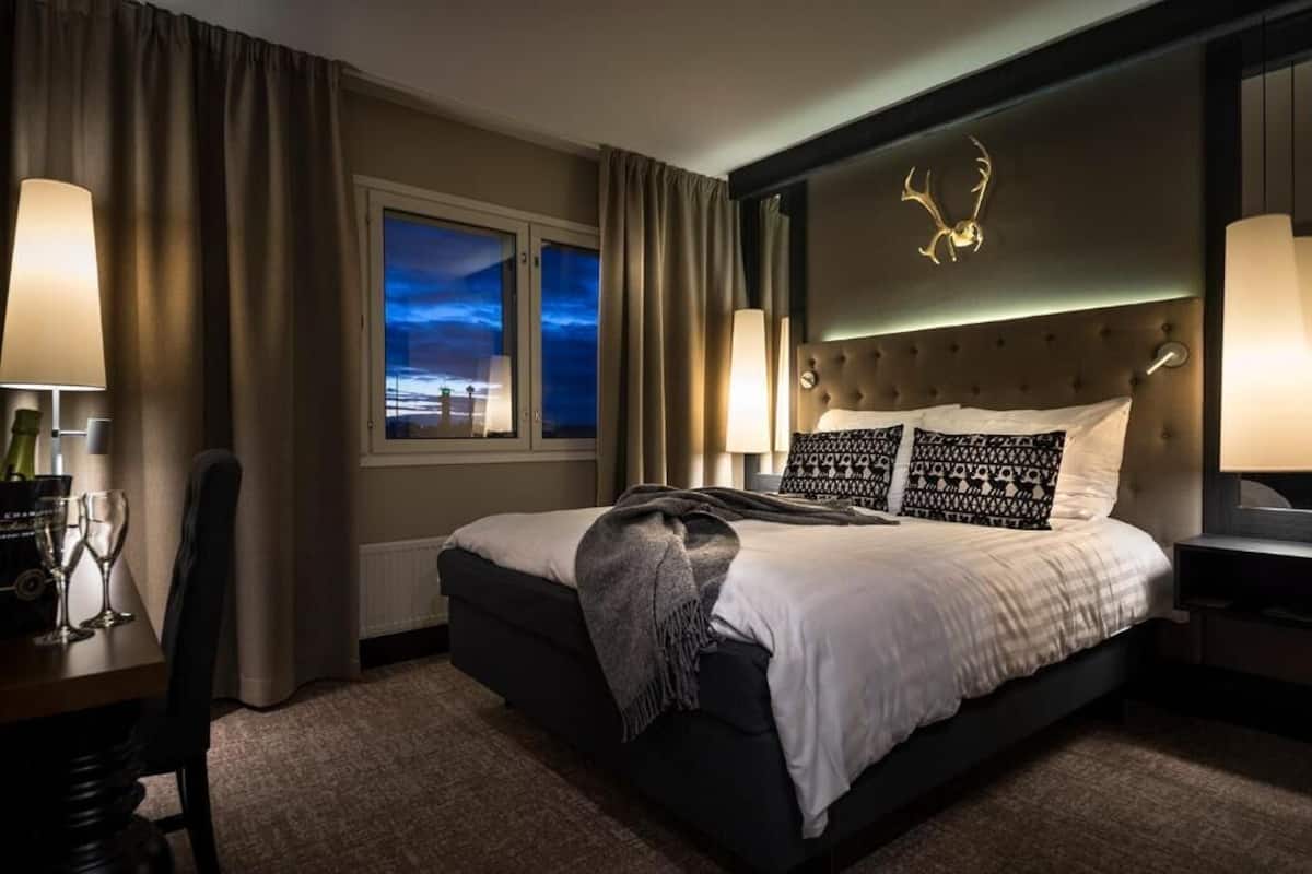 A modern hotel room with a large bed, a view of the cityscape through the window, and a decorative antler on the headboard.