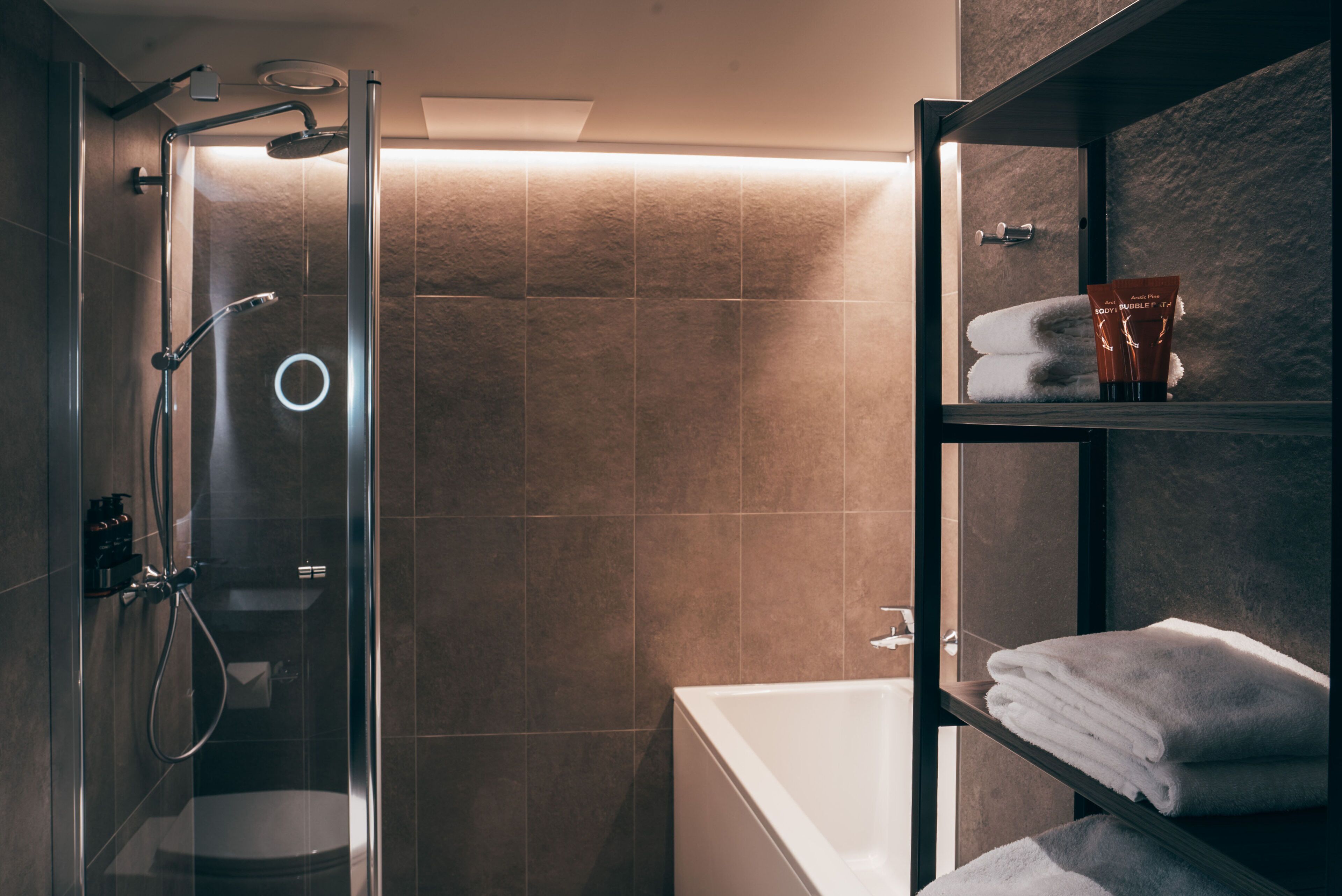 A modern bathroom with a glass-enclosed shower, a white bathtub, and a shelf with folded towels and a toiletry bag.