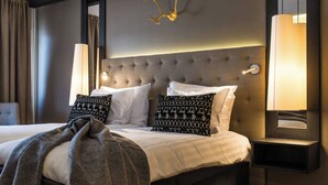 Une chambre d’hôtel avec une tête de lit capitonnée, deux lampes de chevet, un lit avec du linge de lit blanc, des oreillers à motifs et un plaid gris.
