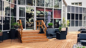 Une terrasse en bois avec des sièges d’extérieur, un escalier menant à une entrée avec porte vitrée et des plantes en pot.
