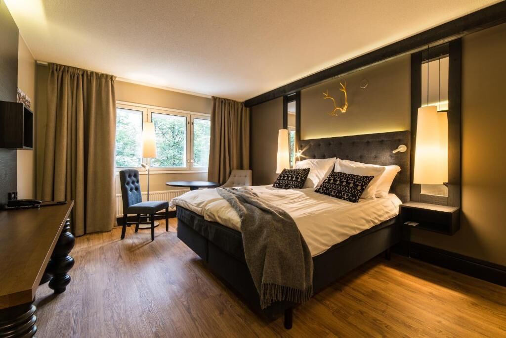 A modern hotel room with a large bed, a desk, and a chair. There is a window with curtains, a lamp, and a wall-mounted TV.