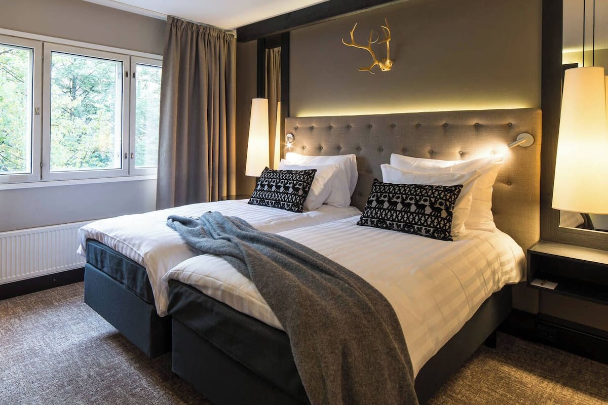 Une chambre d’hôtel avec un grand lit, une tête de lit dotée d’un luminaire, une fenêtre avec des rideaux et un bureau avec une lampe.