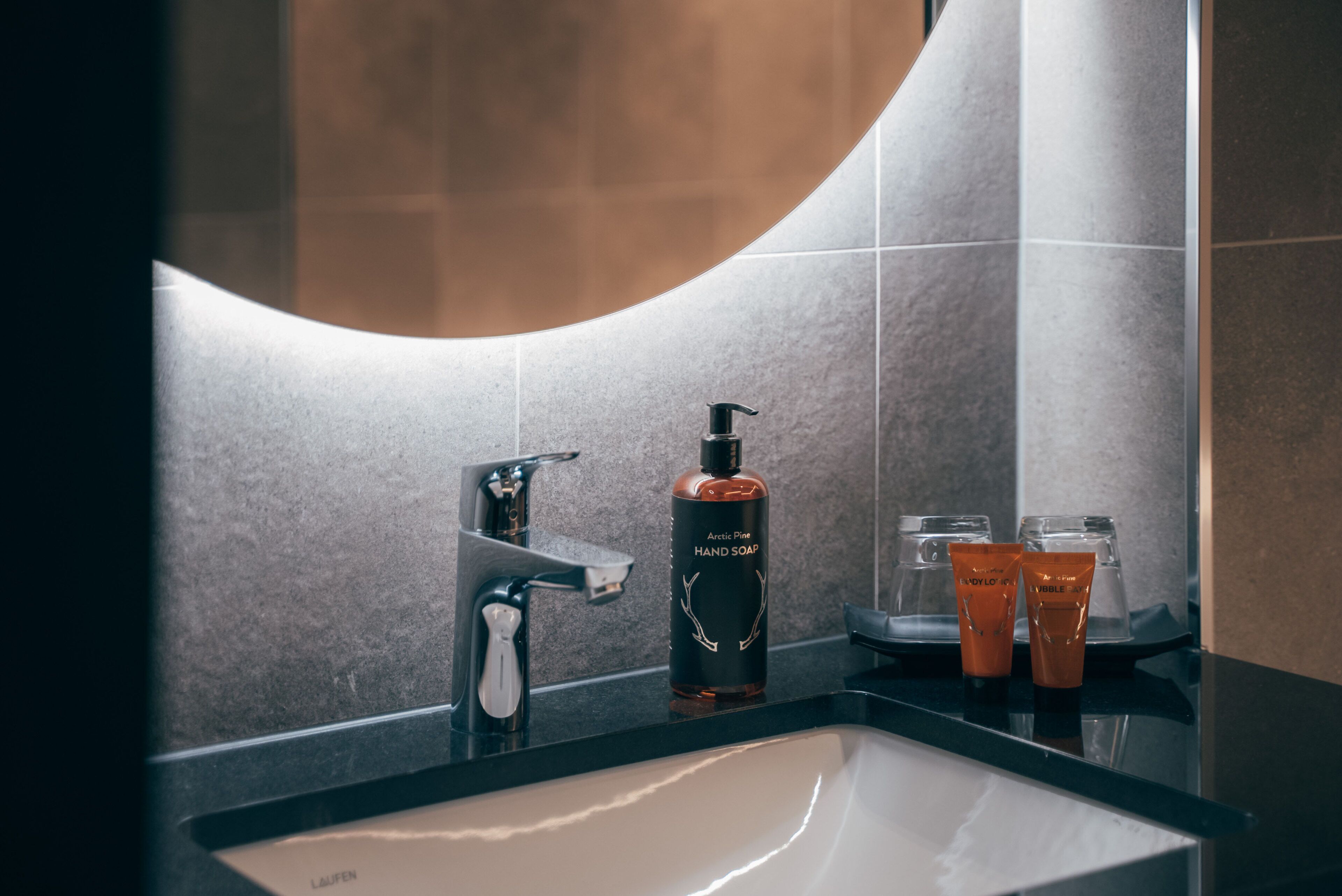 A bathroom with a modern sink, a faucet, a bottle of hand soap, and two orange tubes.