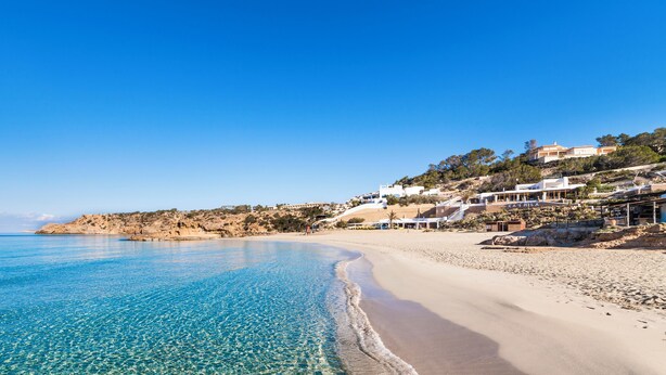 Une plage à l’eau d’un bleu limpide, avec un rivage sablonneux et des bâtiments construits sur une colline.