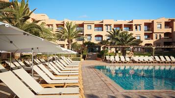 A pool area with sun loungers and umbrellas, a clear sky, and a building with balconies in the background.