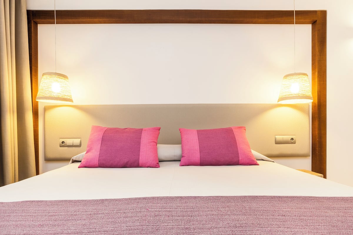 A neatly made bed with two pink pillows, a white headboard, and two wall-mounted lamps.