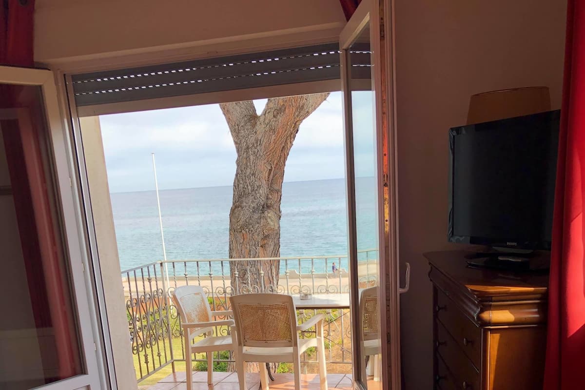Une chambre avec un balcon donnant sur la mer, un téléviseur posé sur un meuble en bois et un lit recouvert d’une couverture rouge.