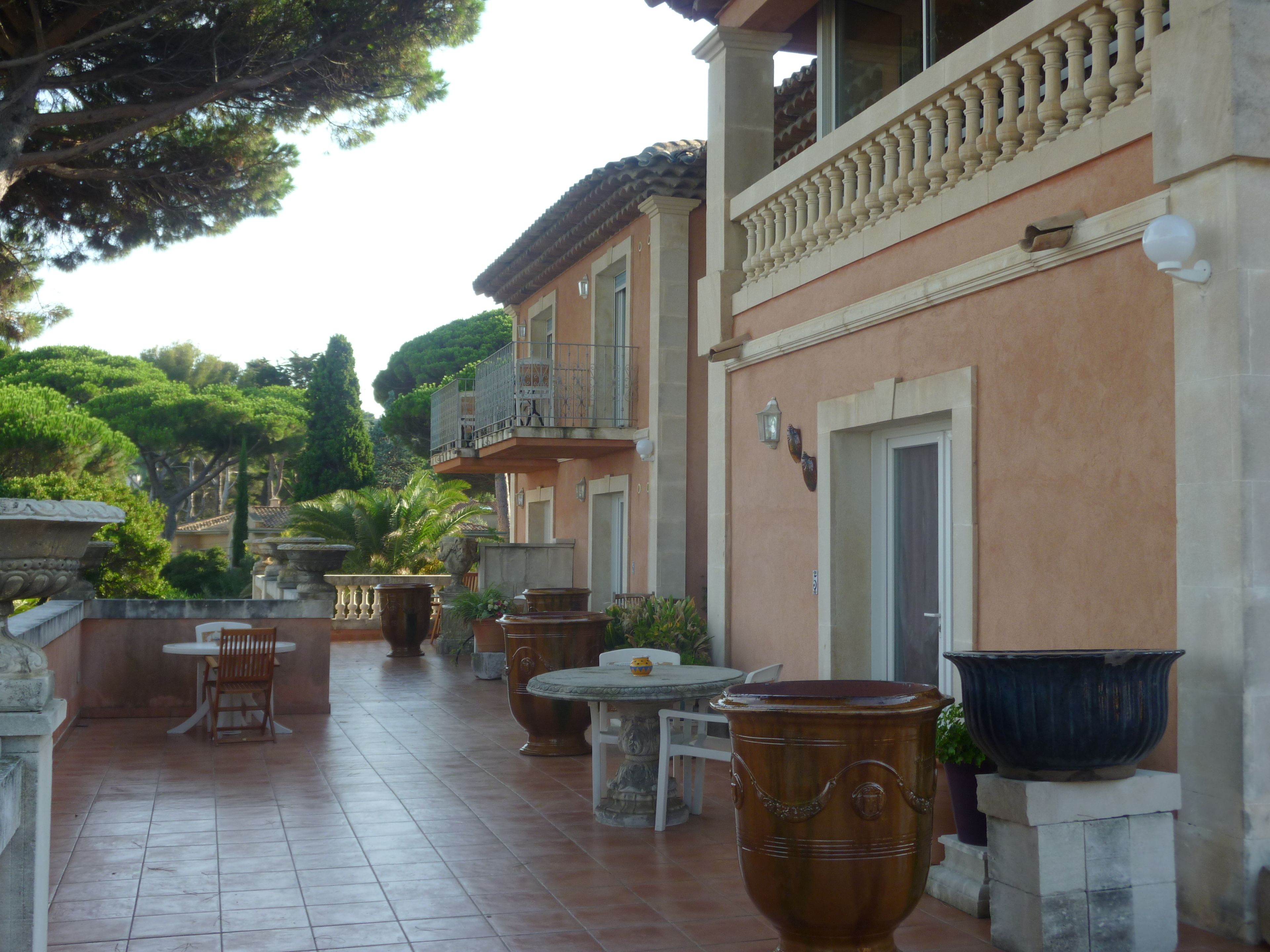 Une cour avec un sol carrelé, des meubles d’extérieur, de grands jardinières et un bâtiment doté d’un balcon.