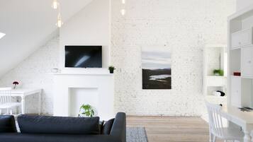 A modern, well-lit room with a white brick wall, a flat-screen TV mounted above a fireplace, a large window with a view, and a white desk with a chair.