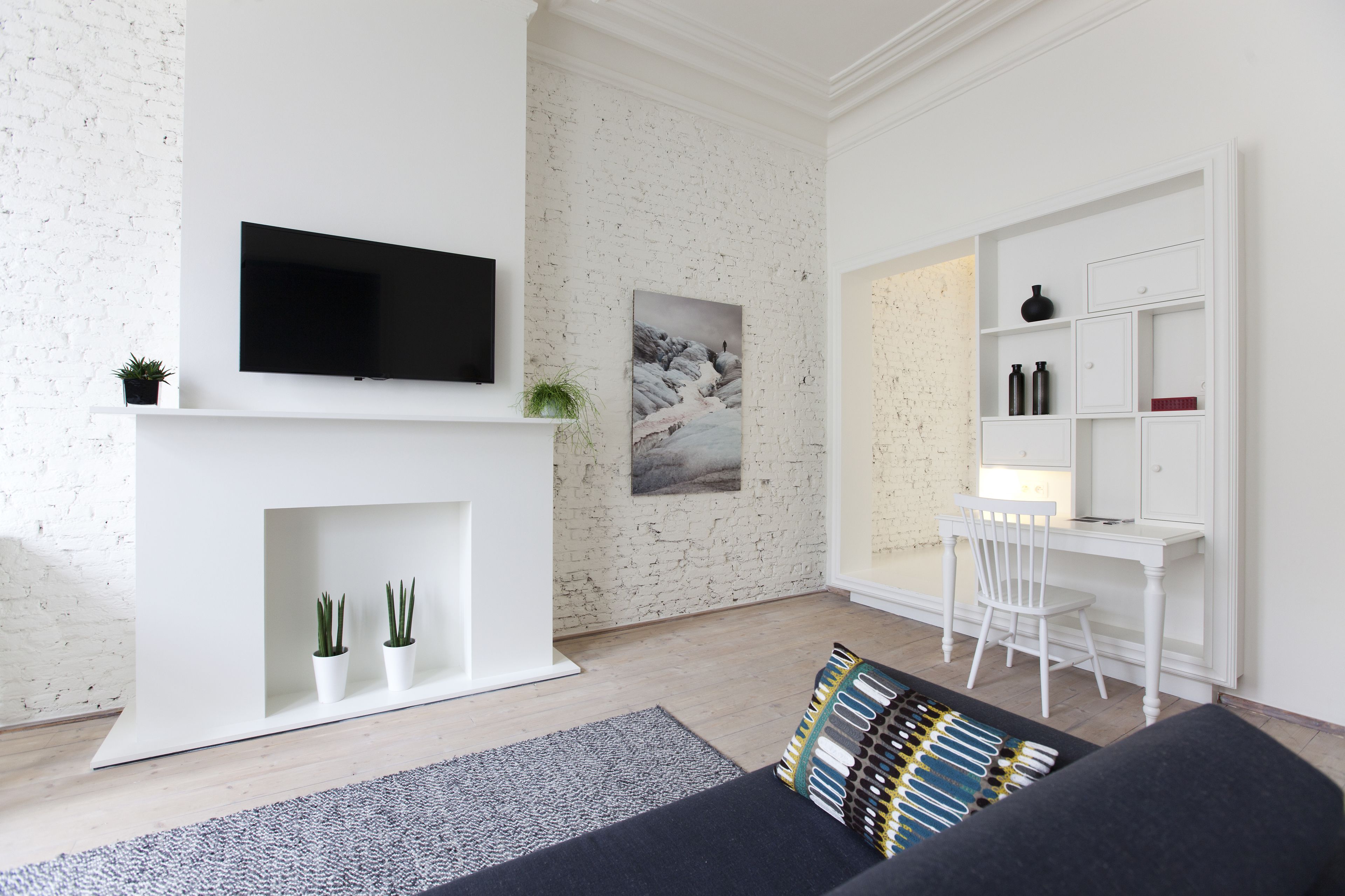 A modern living room with a flat-screen TV mounted above a fireplace, a dining area with a white table and chairs, and a bookshelf with decorative items.