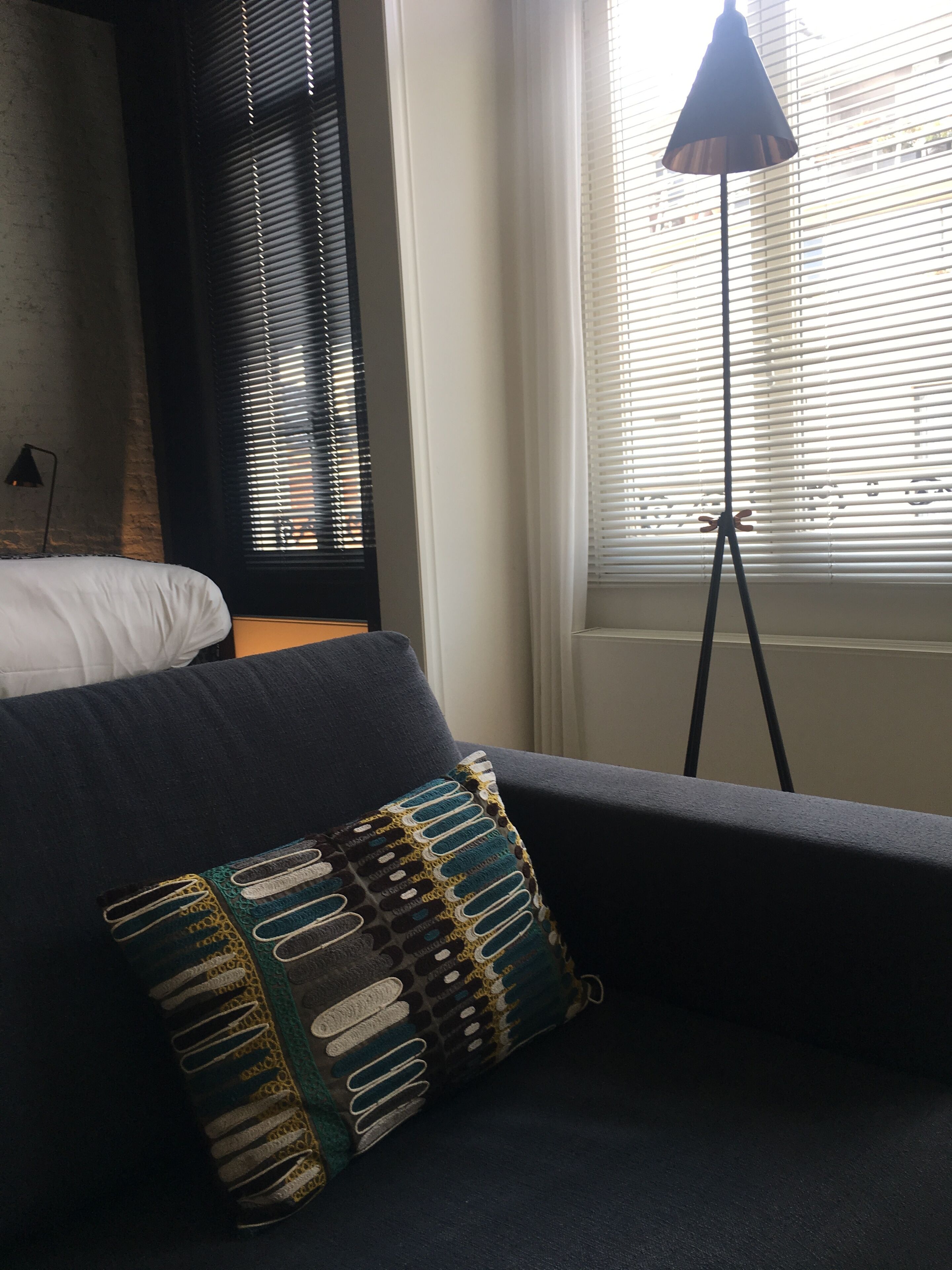 Um quarto com cama, sofá escuro, almofada estampada e luminária de chão perto de uma janela com persianas.